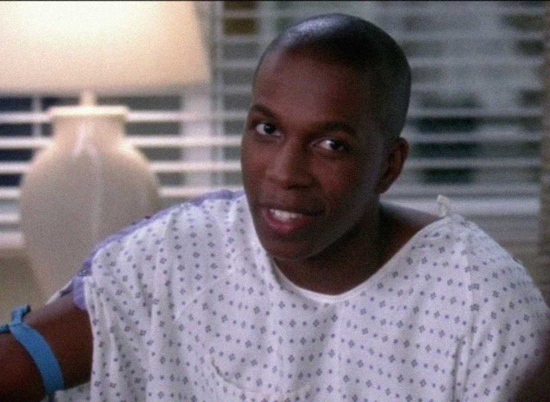 A person wearing a white hospital gown sits with a blue tourniquet on their arm, smiling slightly. A lamp and window blinds are visible in the background.