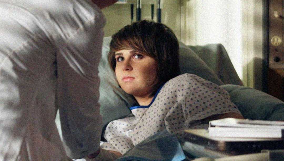 A person with short brown hair lies in a hospital bed, wearing a hospital gown, looking up at someone standing beside the bed who is partially visible. Medical equipment and a tray with books are nearby.
