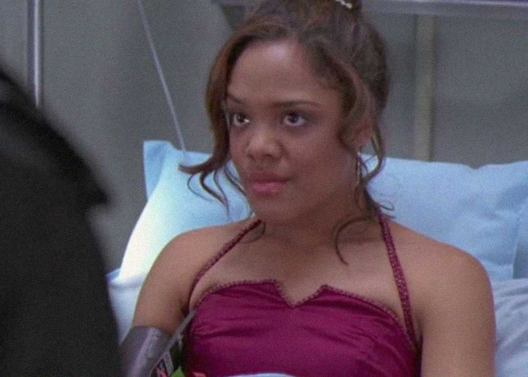 A woman in a hospital bed wears a burgundy strapless dress and looks ahead with a neutral expression. Medical equipment and blue bedding are visible in the background.