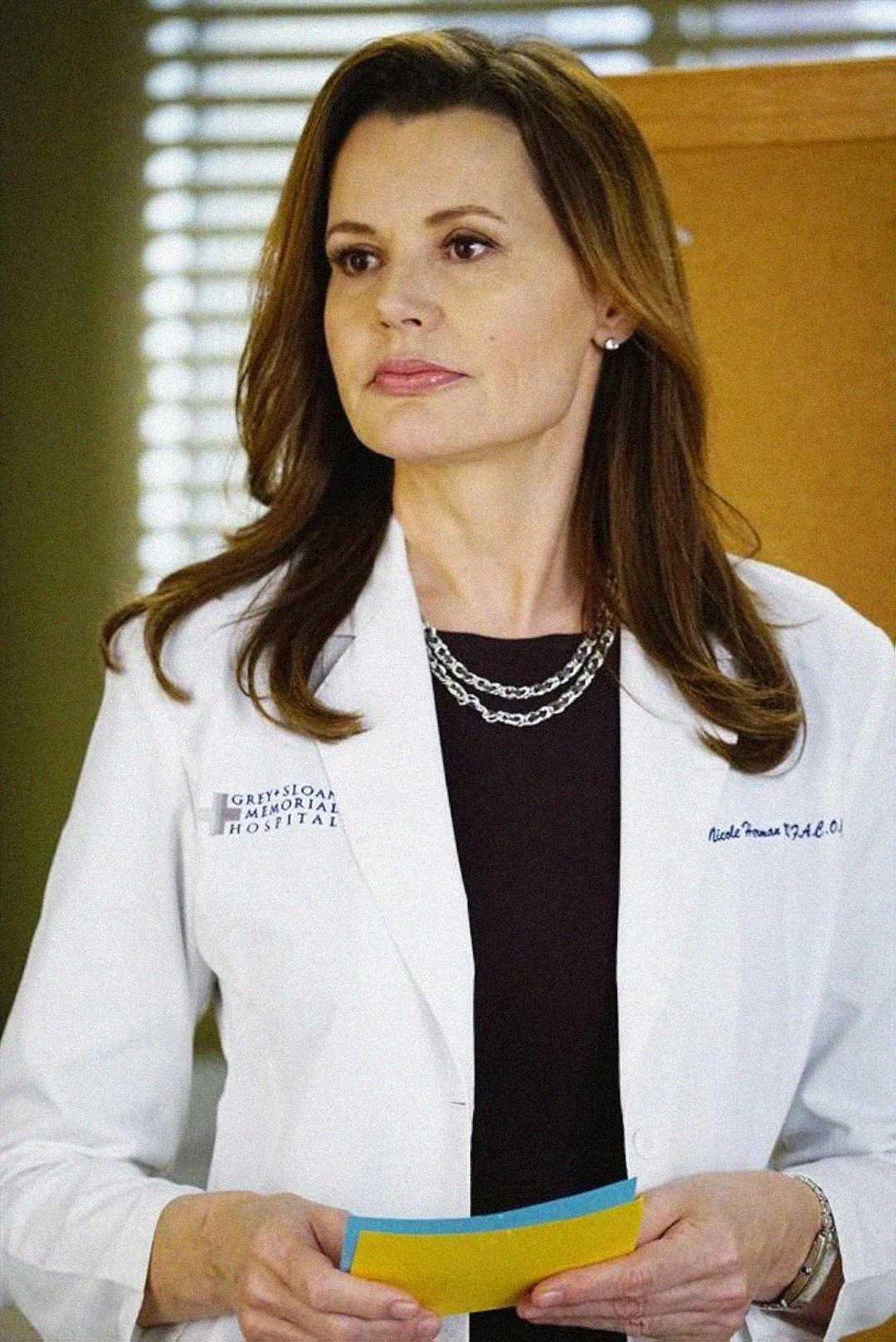 A woman with long brown hair wears a white lab coat and layered necklaces, holding papers. She stands indoors in front of a window with blinds and a corkboard.