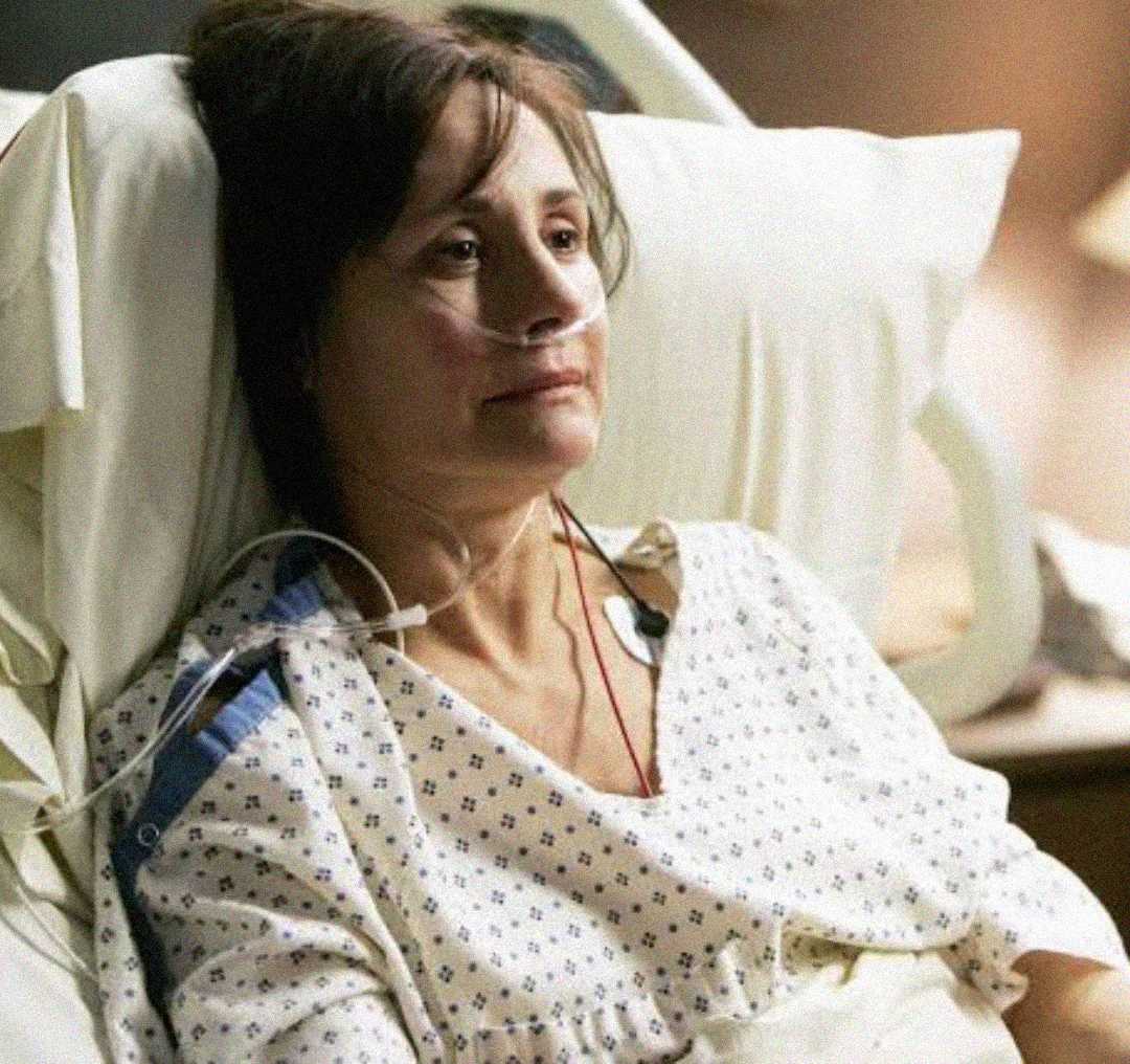 A woman lies in a hospital bed wearing a hospital gown, with a nasal cannula for oxygen and monitoring wires attached to her chest. She appears tired and is looking slightly upward.