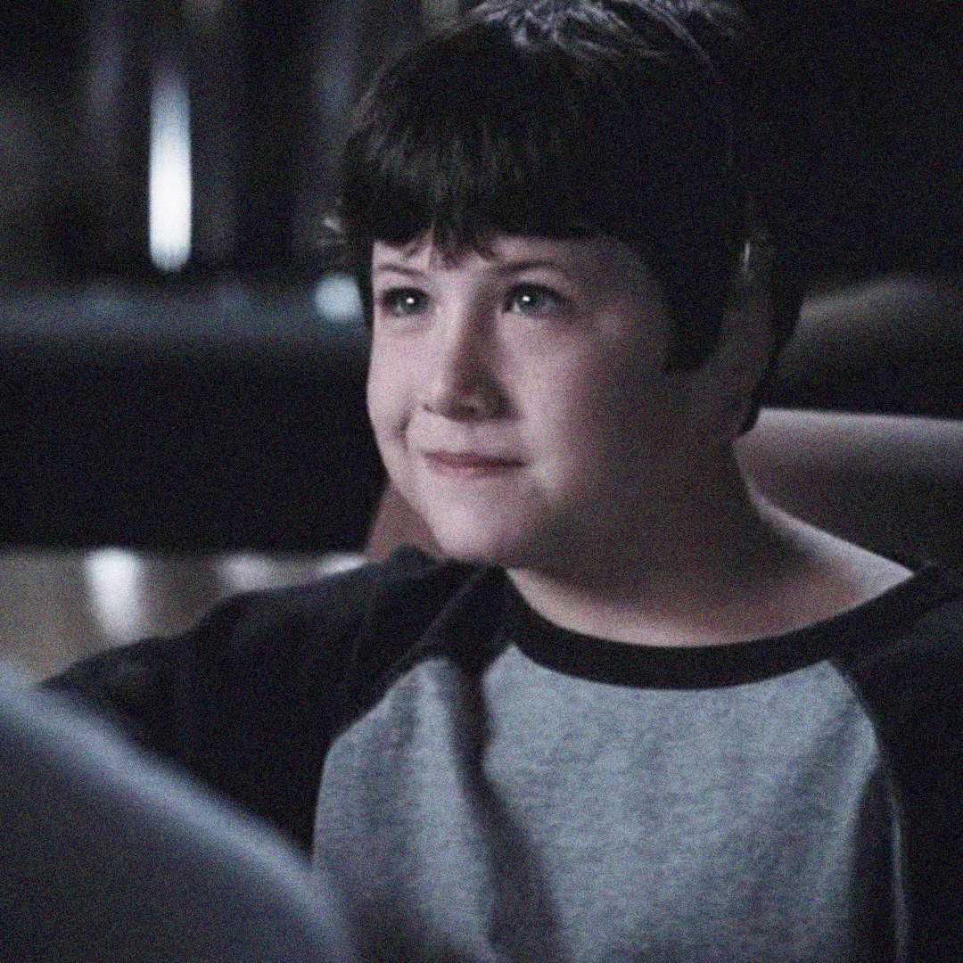 A young boy with short dark hair wearing a black and gray shirt smiles gently while sitting indoors in soft, low light.