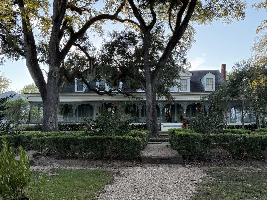A large, two-story house with a wraparound porch, green shutters, and white railings, surrounded by tall trees, hedges, and a gravel pathway leading to the entrance.