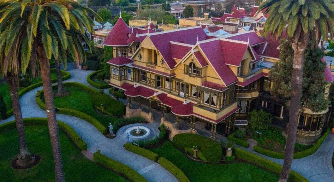 Aerial view of a large Victorian mansion with red roofs, yellow-brown walls, ornate details, manicured lawns, curved pathways, palm trees, and a central fountain in the garden.