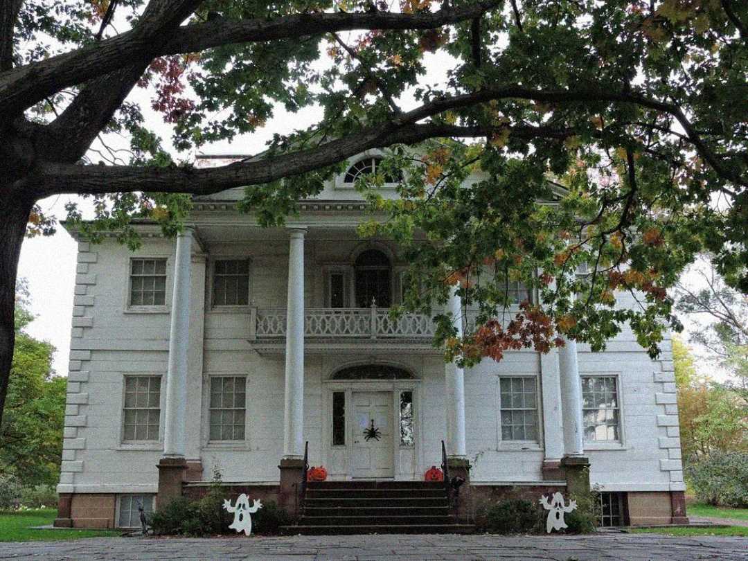 A large white colonial-style house with tall columns, a balcony, and double front doors. Autumn leaves hang from a tree above, and two white ghost decorations stand near the steps.
