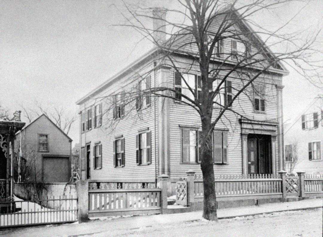 Black-and-white photo of a two-story, wood-frame house with shutters, a front porch, and a leafless tree in front. A picket fence runs along the sidewalk, and another house is visible in the background.