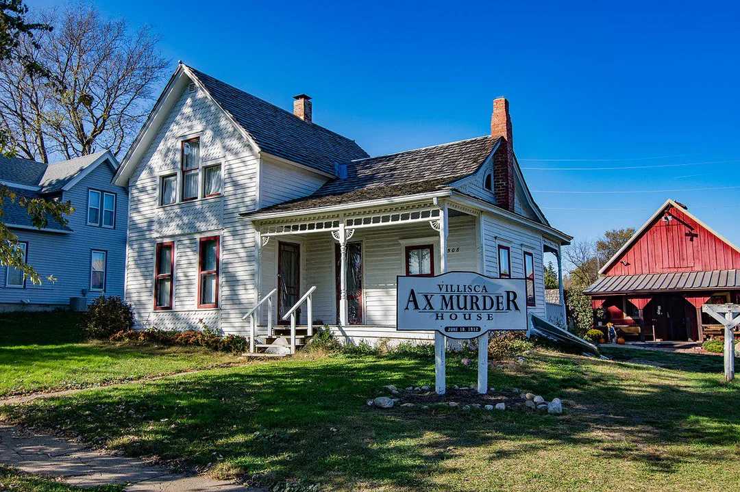 A white, two-story historic house with a front porch and a sign reading "Villisca Ax Murder House" stands beside a red barn under a clear blue sky.