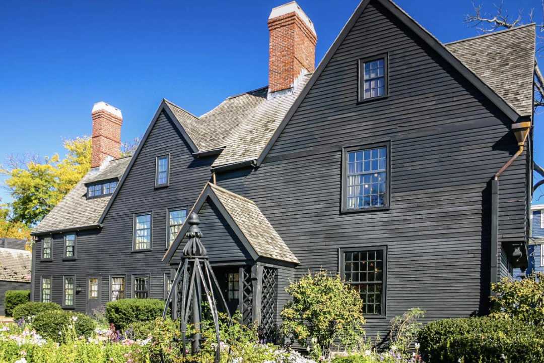 A historic dark wood house with steep gabled roofs, large brick chimneys, and a garden with green bushes and flowering plants under a clear blue sky.