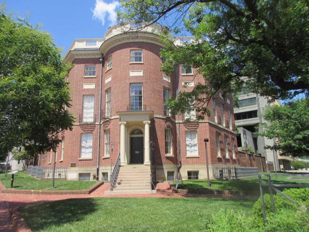 A historic red brick building with white-trimmed windows, arched doorway, and stairs leading to the entrance, surrounded by green trees and grass on a sunny day.