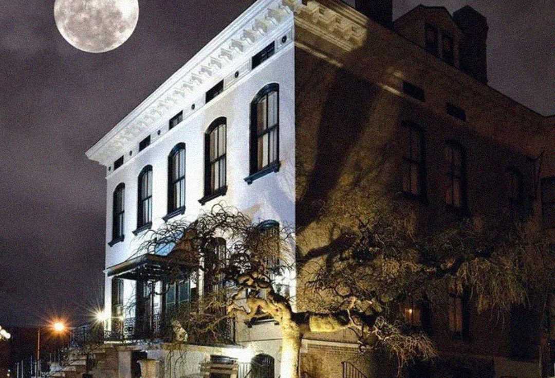 A large, old building with arched windows is dramatically lit at night. A gnarled tree stands in front, and a bright full moon shines in a cloudy sky, creating a mysterious atmosphere.
