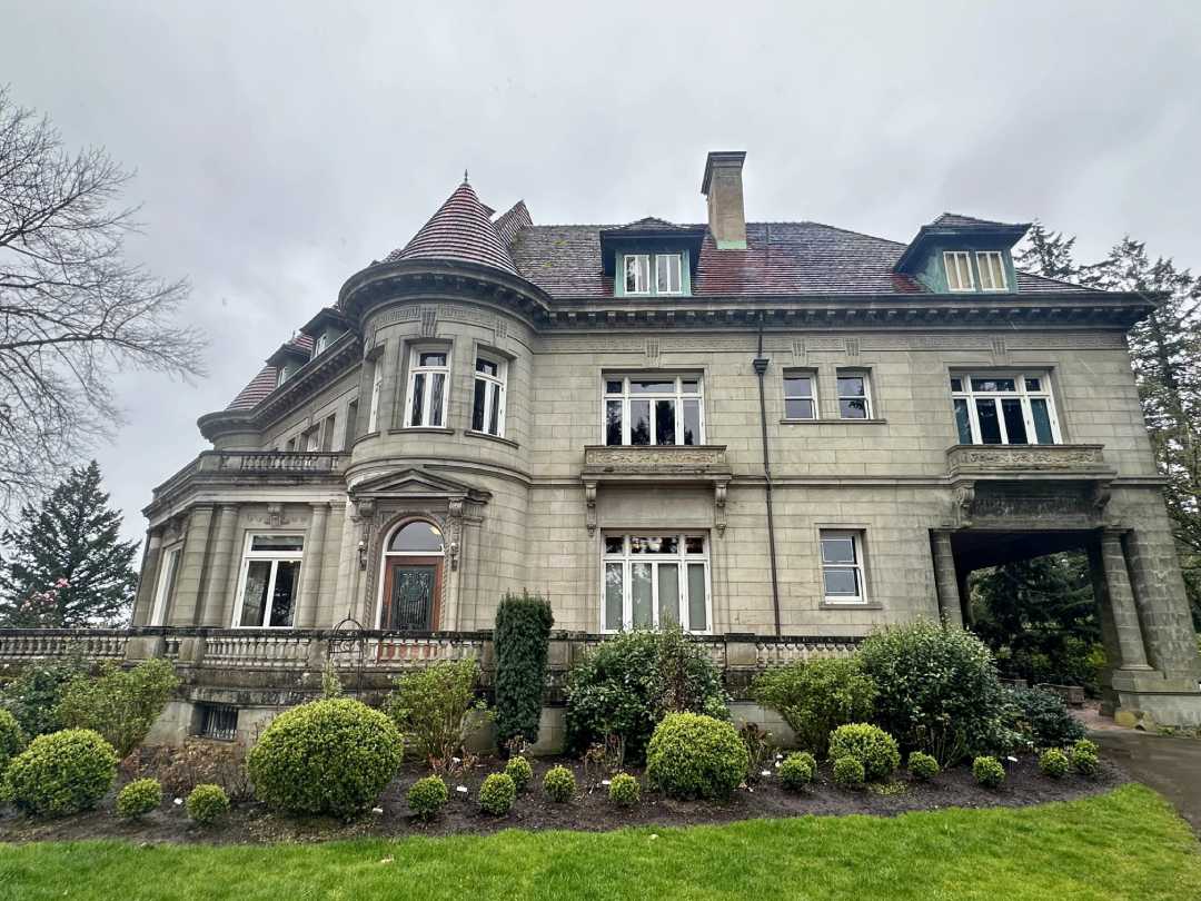 A large, historic stone mansion with ornate windows, a red-tiled roof, and manicured bushes in front, set against a cloudy sky.