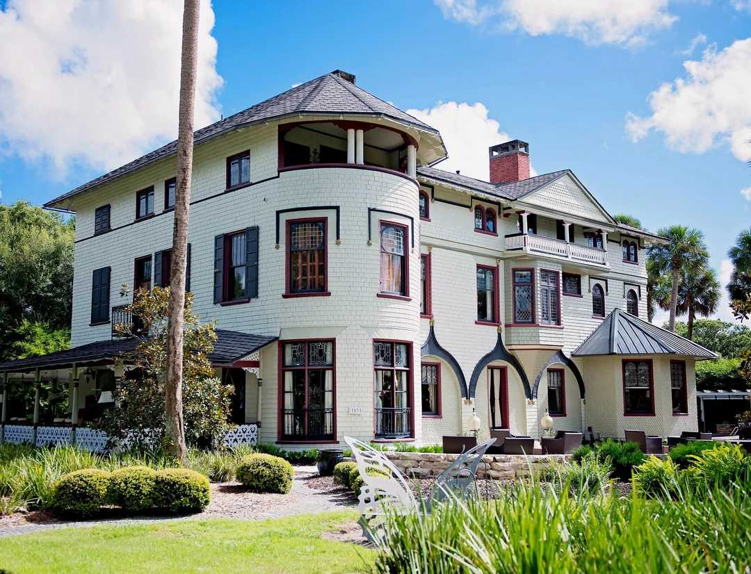 Large, historic three-story house with cream-colored siding, dark trim, and a round turret, surrounded by green lawn, palm trees, and manicured bushes under a bright blue sky with scattered clouds.