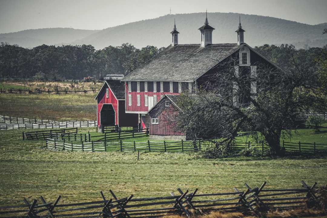 A large red barn with white trim sits behind a split-rail wooden fence, surrounded by grass and trees, with rolling hills and mountains in the background under a hazy sky.