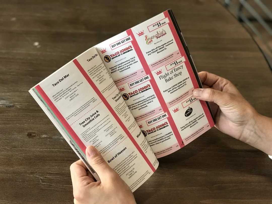 A person holds a booklet open to pages filled with coupons and advertisements for local businesses, including restaurants and bakeries, on a wooden table.