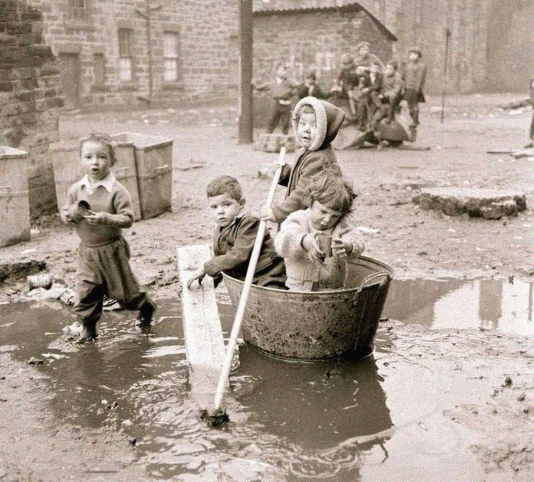 Four young children play in a muddy street; two sit in a large metal tub with sticks, while one stands beside it holding a plank and another walks nearby. Old brick buildings and more children are in the background.
