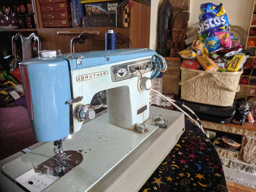 A vintage blue and white Brother sewing machine sits on a table. In the background, there is a bowl filled with colorful snack bags and shelves with various items, including books and decorations.