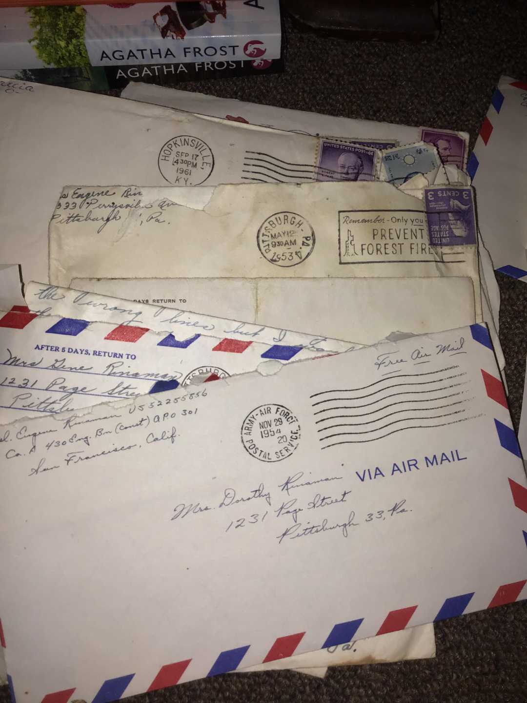 A few vintage envelopes, some labeled "Via Air Mail," with handwritten addresses and postage stamps are spread out on a surface. Part of an Agatha Frost book is visible in the background.