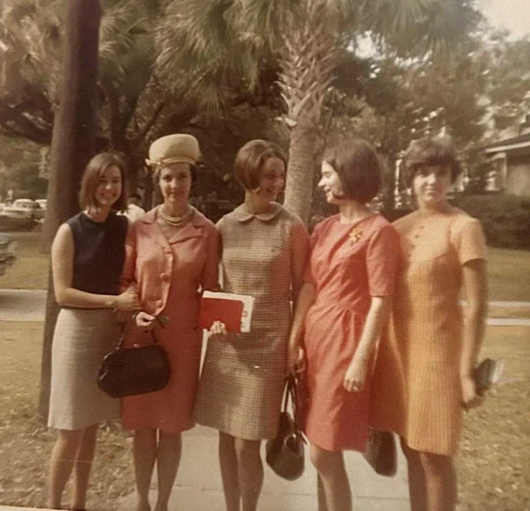 Five women stand outdoors on a sidewalk lined with palm trees, wearing 1960s-style dresses in various colors and smiling at each other. The background includes grass, trees, and residential buildings.