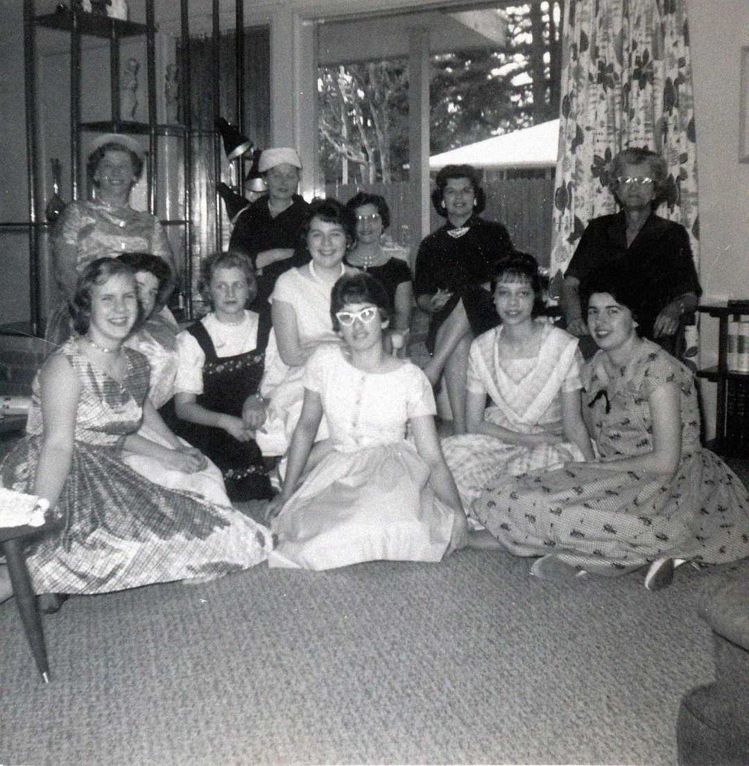 A black-and-white photo of eleven women and girls, some sitting on the floor and others on chairs, in a living room decorated with curtains, a bookshelf, and a display shelf. Most are smiling and dressed in 1950s-style clothing.