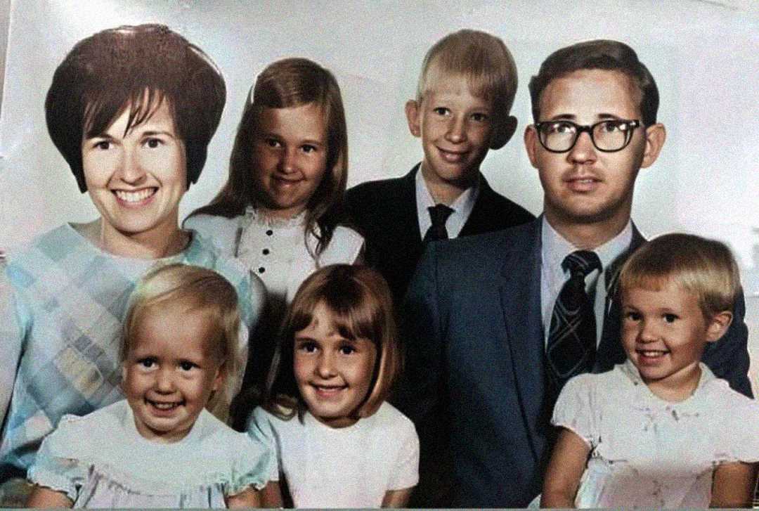 A vintage family portrait of two adults and four children, all smiling. The man wears glasses and a suit; the woman has short dark hair and wears a patterned dress. The children, three girls and one boy, are dressed in light-colored clothes.