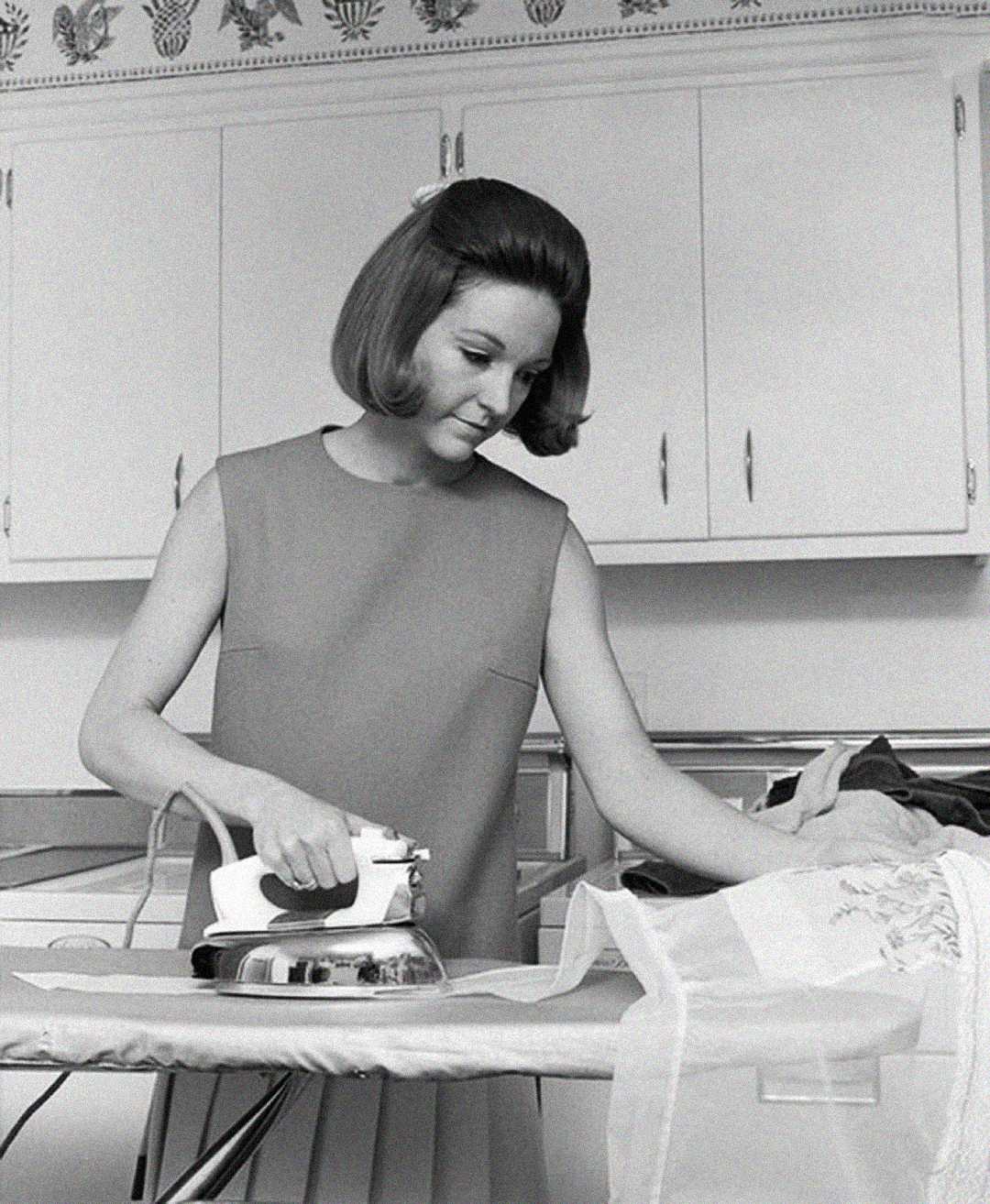 A woman with a bob hairstyle wears a sleeveless dress and irons clothing on an ironing board in a vintage kitchen with white cabinets.