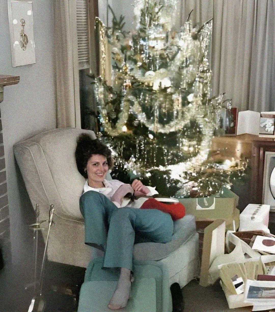 A woman sits smiling on a chair in a cozy living room, surrounded by opened gifts and wrapping paper, with a decorated, lit Christmas tree in the background.