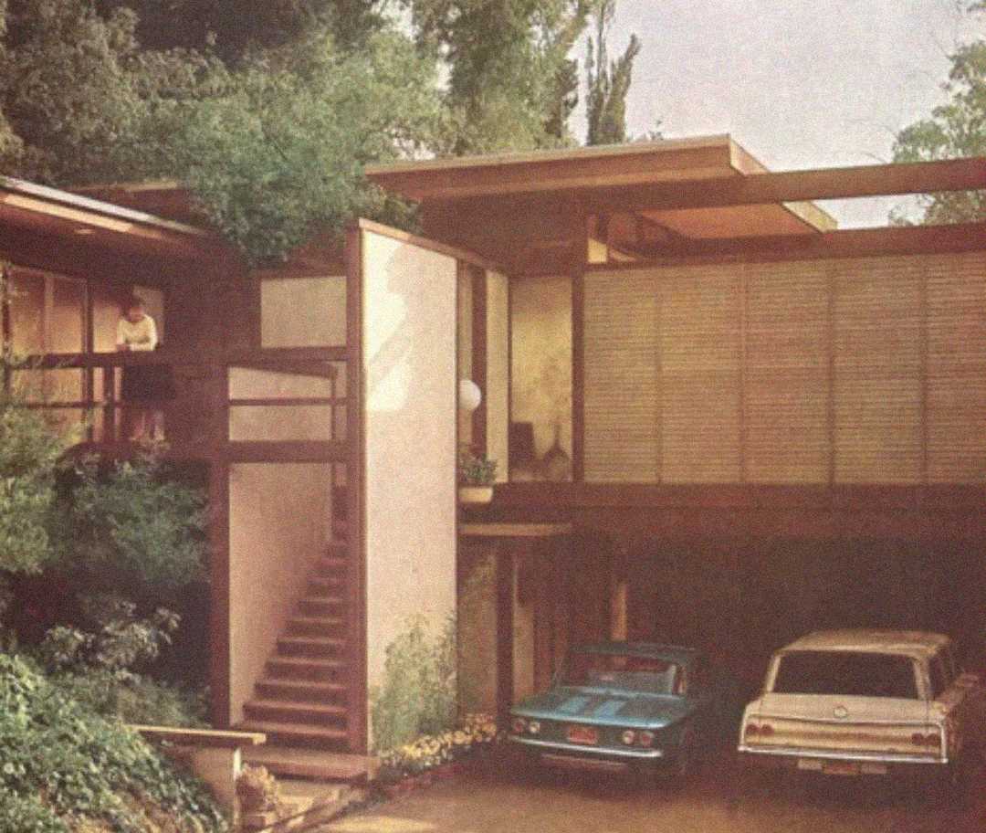A modern, two-story house with large windows, wooden accents, and a carport. Two vintage cars are parked under the house, and a person stands on the upper balcony near lush greenery.