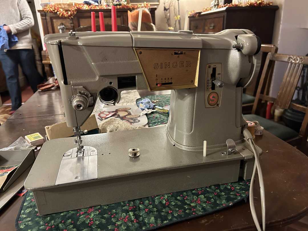 A vintage Singer sewing machine sits on a table with a green floral mat, surrounded by sewing supplies. In the background, furniture and holiday decorations are visible, with a person standing to the left.