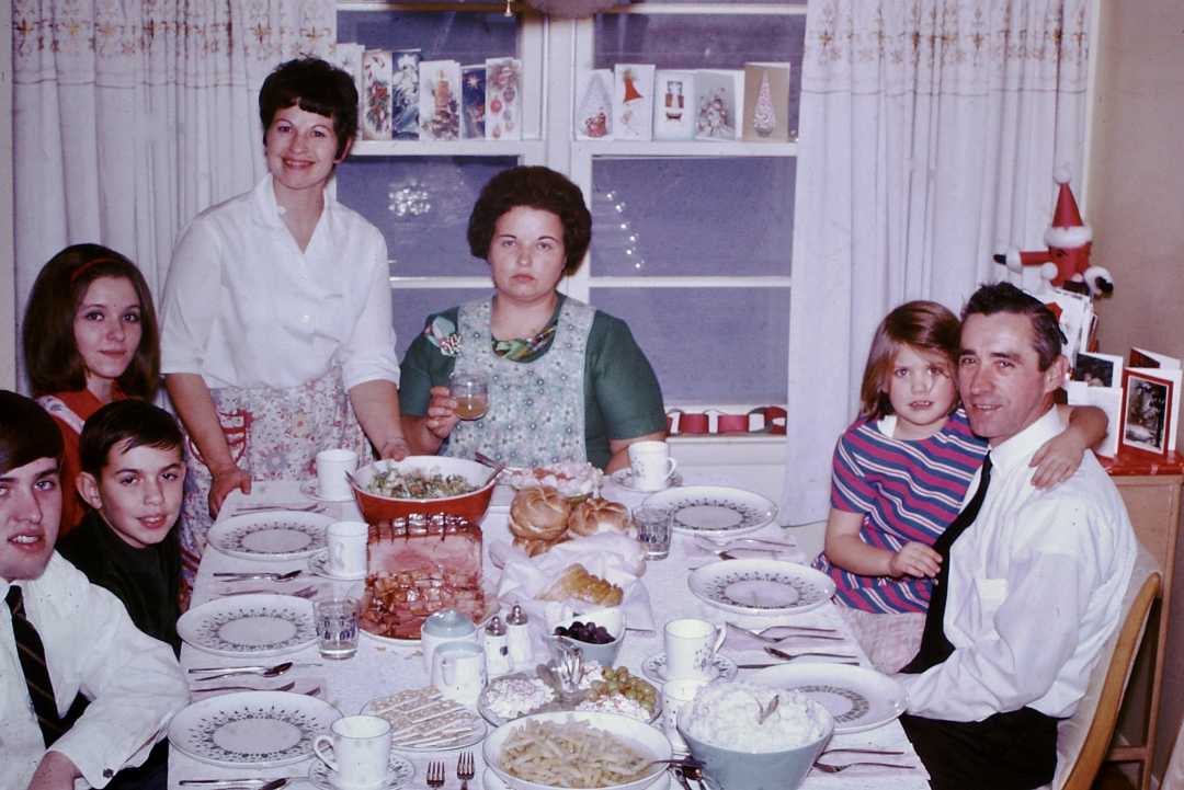 A family of seven sits around a dining table set with food, including bread, salad, and desserts. The room has white curtains, holiday cards on the window, and everyone is dressed nicely, smiling or posing for the photo.