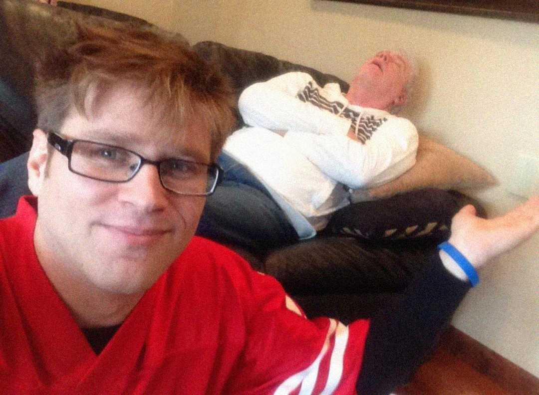 A man in glasses takes a selfie while smiling, wearing a red sports jersey. In the background, an older man lies on a couch with his arms crossed, appearing to be sleeping or laughing with his head back.