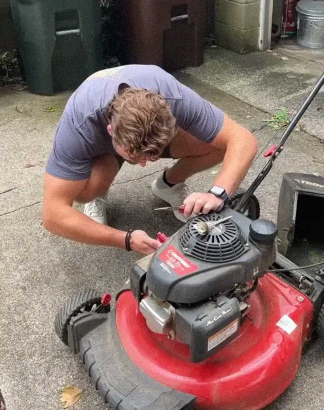 A person crouches on a concrete driveway, using a tool to repair or adjust the engine of a red lawn mower. Two green trash bins and a brown container are visible in the background.