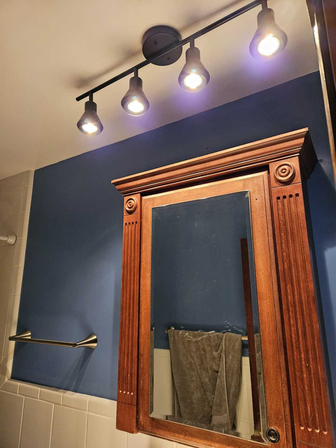 A wooden bathroom medicine cabinet with a mirror is mounted on a blue wall. Above it, four ceiling spotlights are turned on. A towel hangs on a metal bar to the left. White tiles cover part of the wall.
