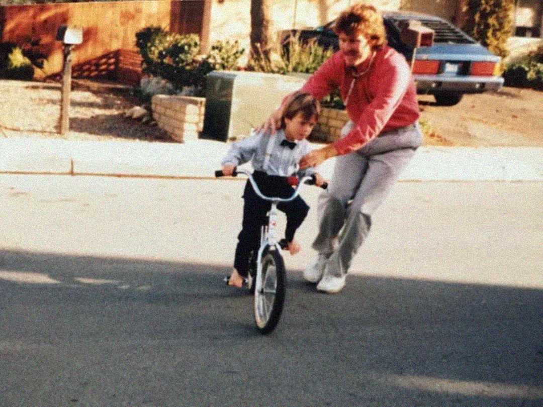 An adult helps a young child ride a bicycle on a suburban street. The child is focused on riding while the adult supports them from behind. Houses, a parked car, and trees are visible in the background.