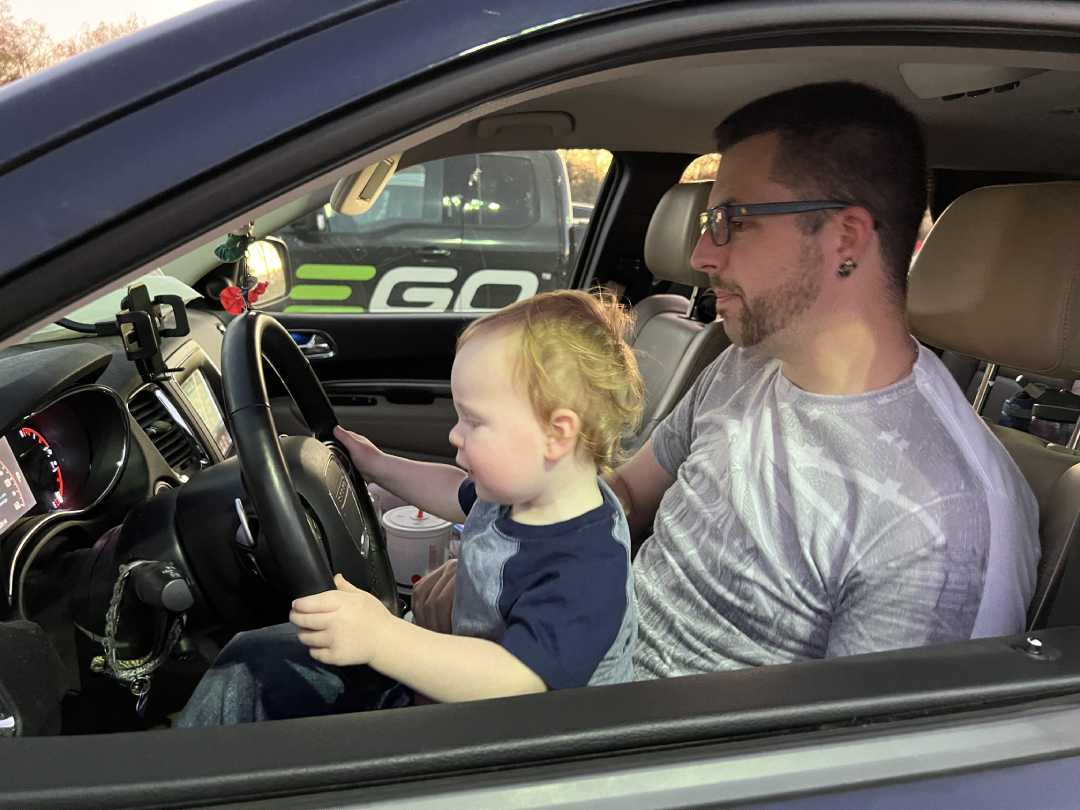 A man sits in the driver’s seat of a car with a young child on his lap, both looking at the steering wheel. The man wears glasses and a t-shirt; the child appears to be playing with the controls.