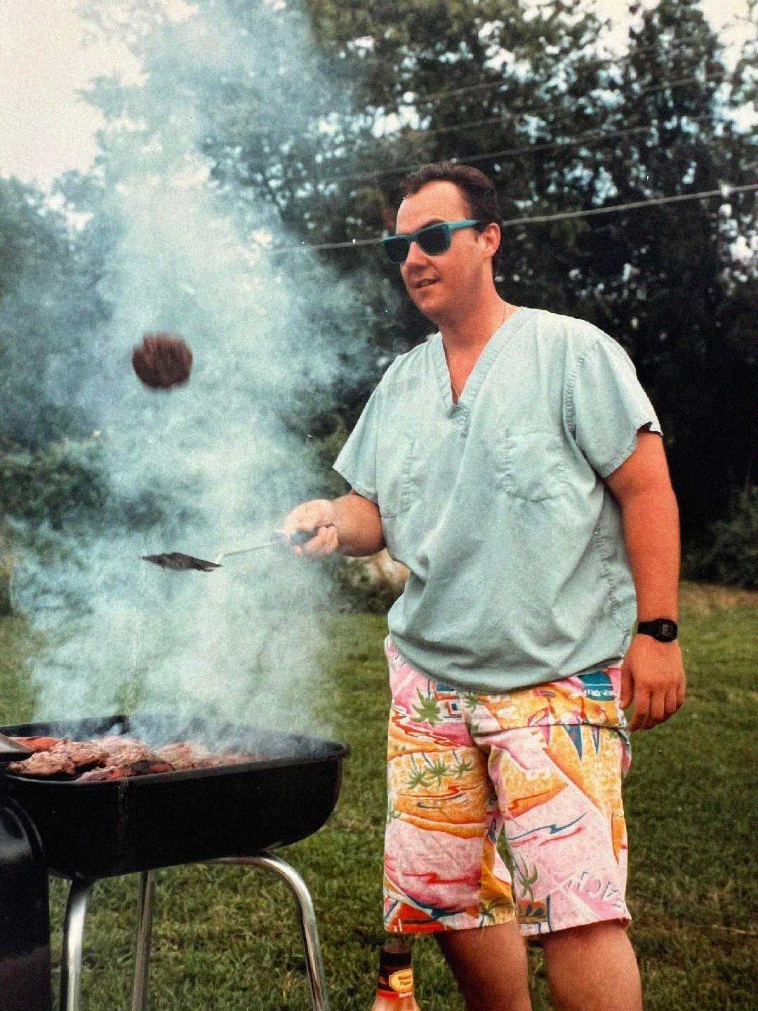 A man in scrubs and colorful shorts flips a burger on a smoky outdoor grill, wearing sunglasses and standing on green grass with trees and power lines in the background.