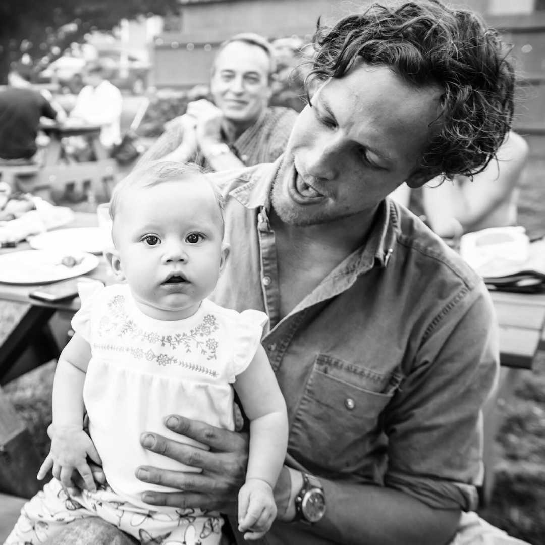 A man holds a baby on his lap at an outdoor gathering. The baby looks at the camera while the man looks at the baby and smiles. Other people are visible in the background, sitting at picnic tables.