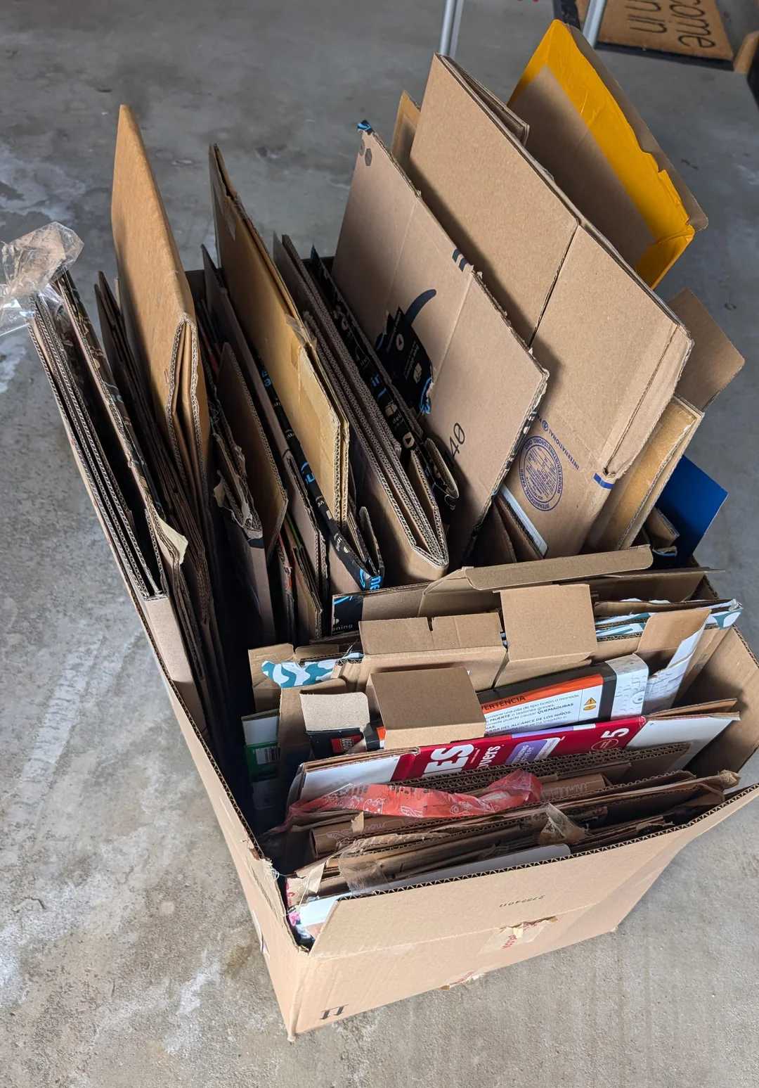 A large cardboard box filled with flattened cardboard pieces and packaging materials, organized upright in the box and placed on a concrete floor.