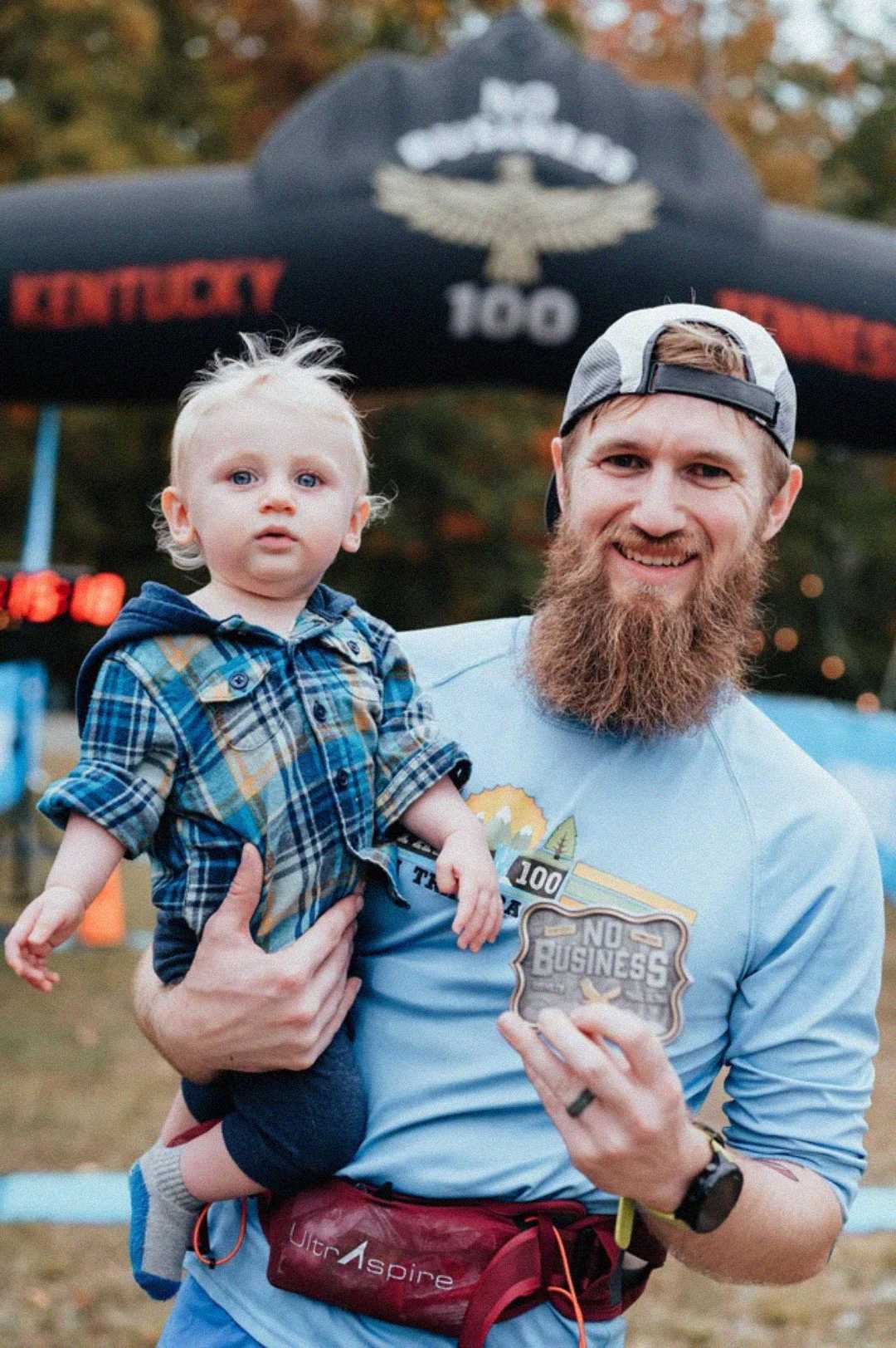 A smiling man with a long beard holds a toddler in his arms at an outdoor event. The man wears a race bib and a running shirt, with a "No Business 100" sign and finish line arch visible in the background.