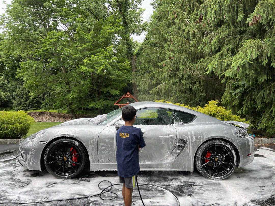 A child stands outside washing a silver sports car covered in soap suds, surrounded by lush green trees and bushes on a cloudy day.