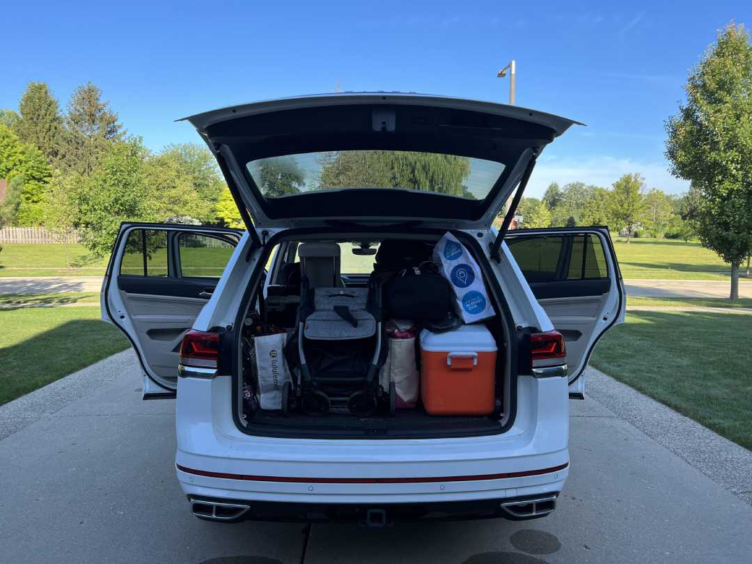 A white SUV with its trunk and rear doors open is parked on a driveway. The trunk is packed with luggage, a cooler, rolls of paper towels, and other travel items. Green trees and grass are visible in the background.
