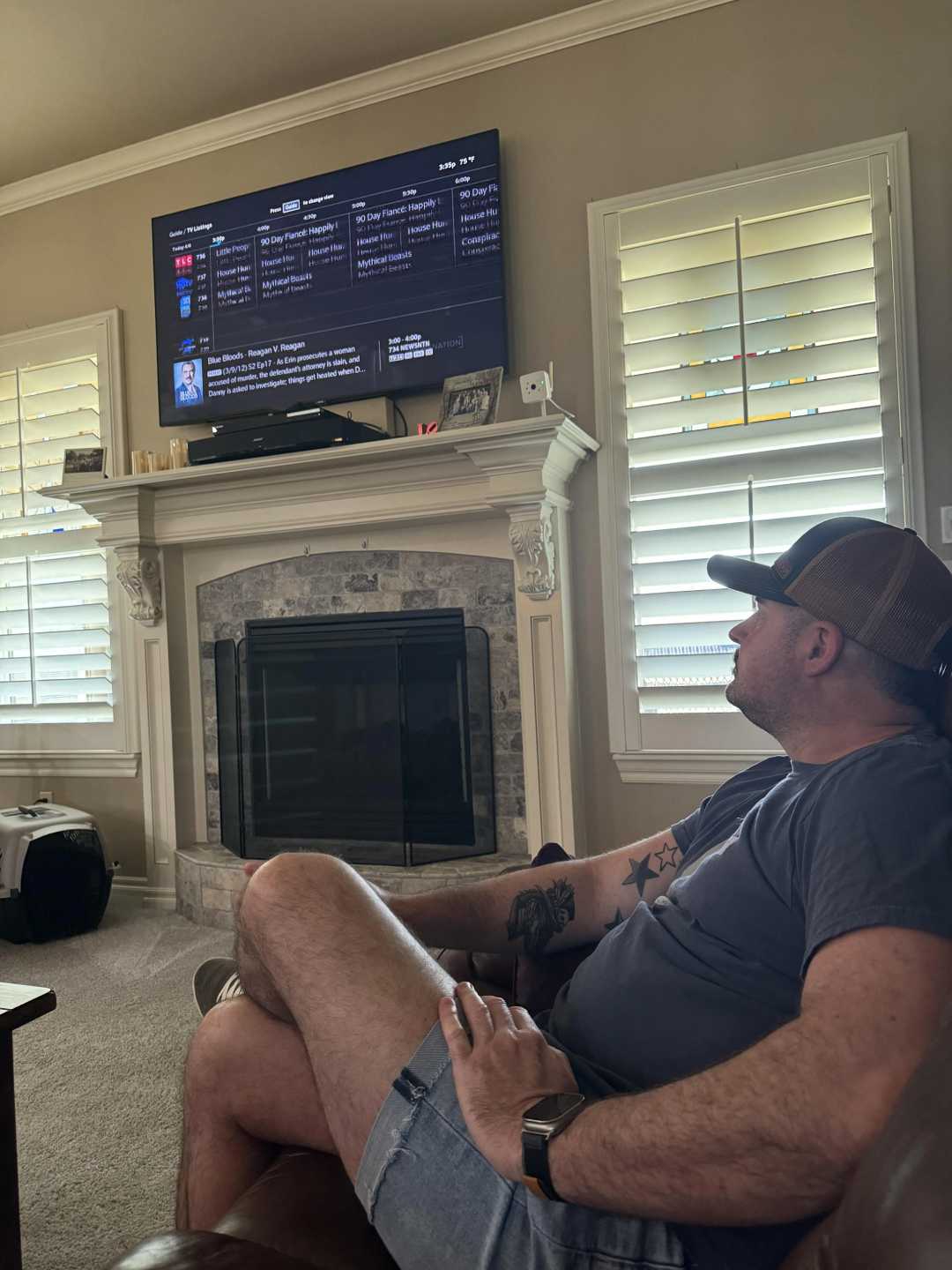 A man in a cap and shorts sits on a couch, watching a wall-mounted TV above a fireplace in a living room with shuttered windows and neutral decor.