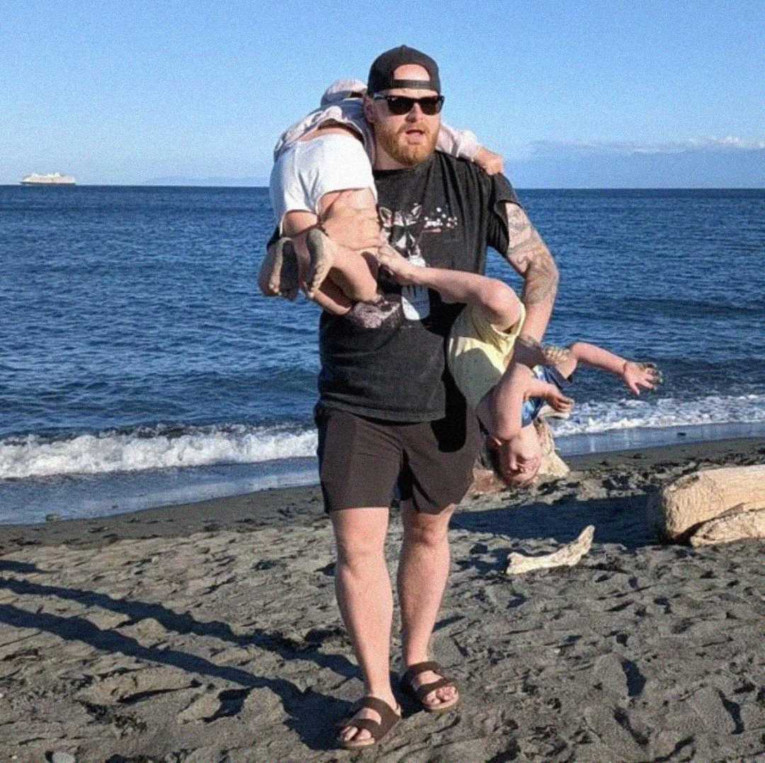 A man in shorts and a cap stands on a sandy beach, holding two young children—one under each arm—both hanging upside down as they playfully dangle. The ocean and blue sky are in the background.