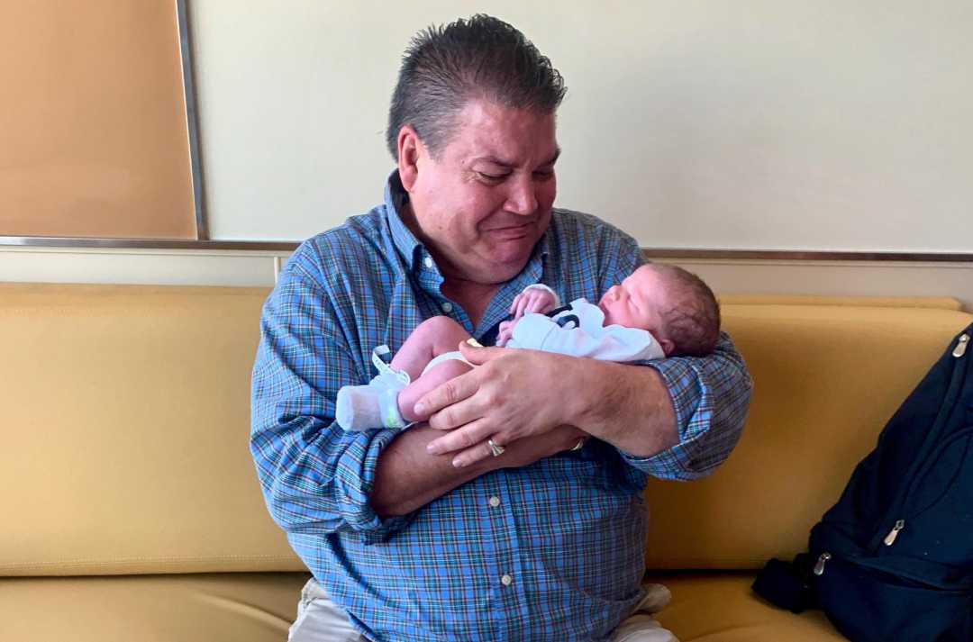 A man wearing a blue plaid shirt sits on a yellow couch, cradling a newborn baby dressed in light blue clothing and smiling down at the baby. A dark bag rests beside him.