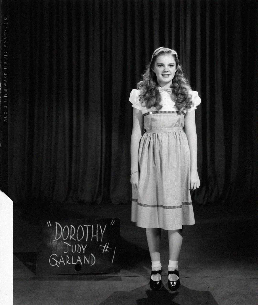 A young woman with curly hair stands in front of a curtain, wearing a vintage dress and Mary Jane shoes. At her feet, a board reads: '"DOROTHY" JUDY GARLAND #1.' The photo is in black and white.