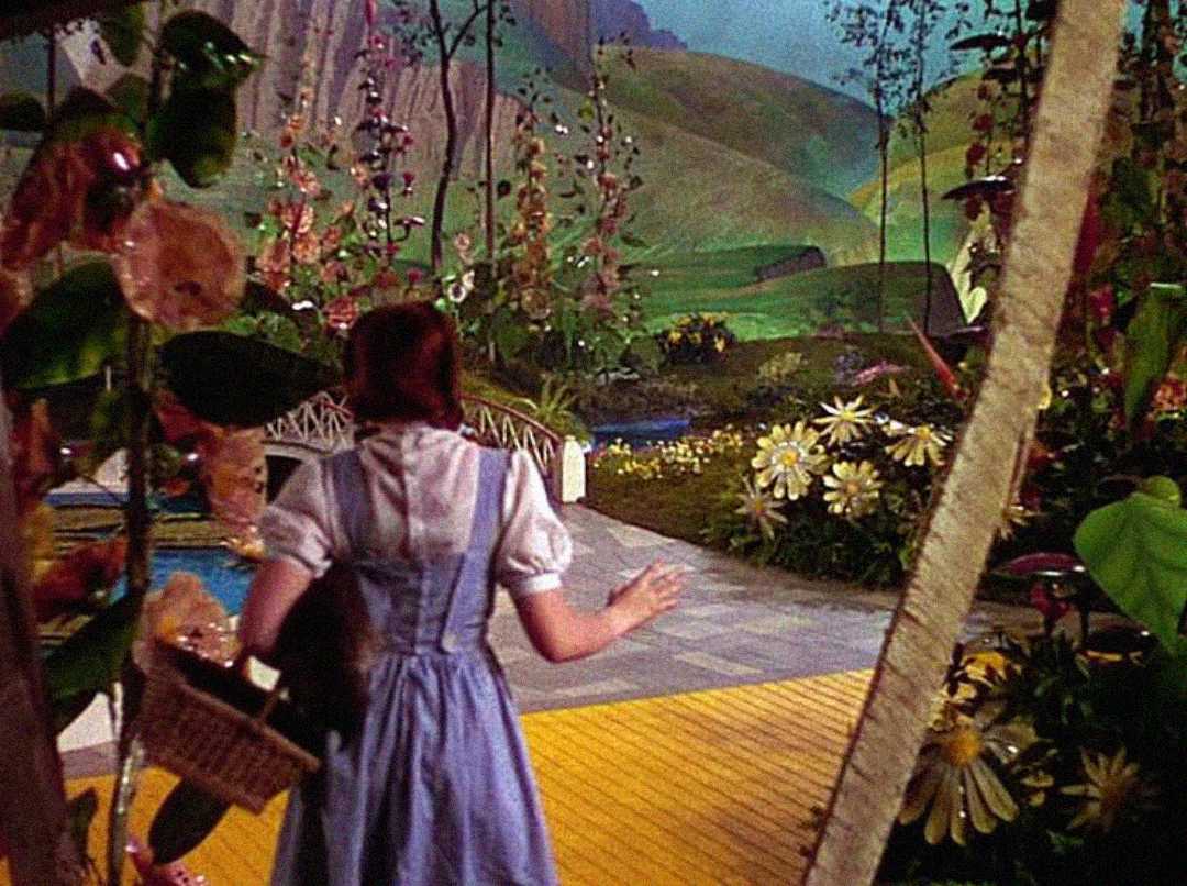 A girl in a blue dress stands at the start of a yellow brick road, surrounded by vibrant flowers and greenery, looking toward a scenic, colorful landscape with hills in the distance.