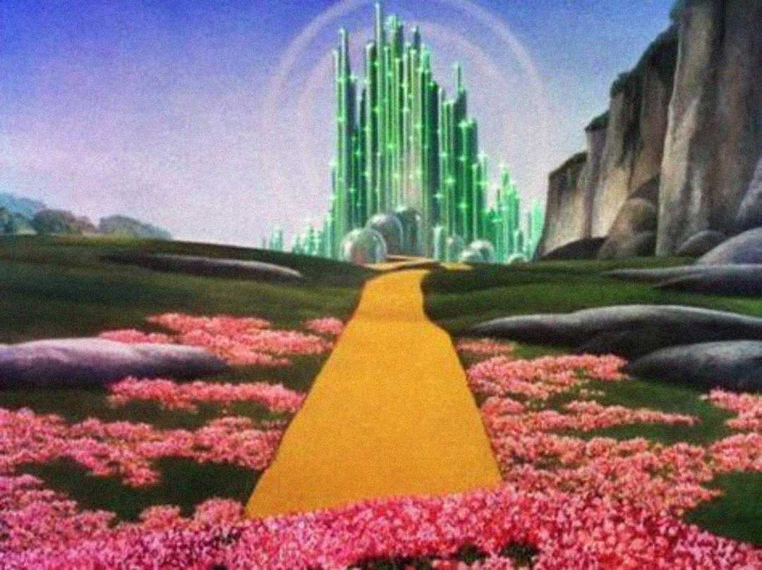 A yellow brick road winds through a field of pink flowers toward a glowing, green, crystal-like city beneath a blue sky with distant cliffs on the right.