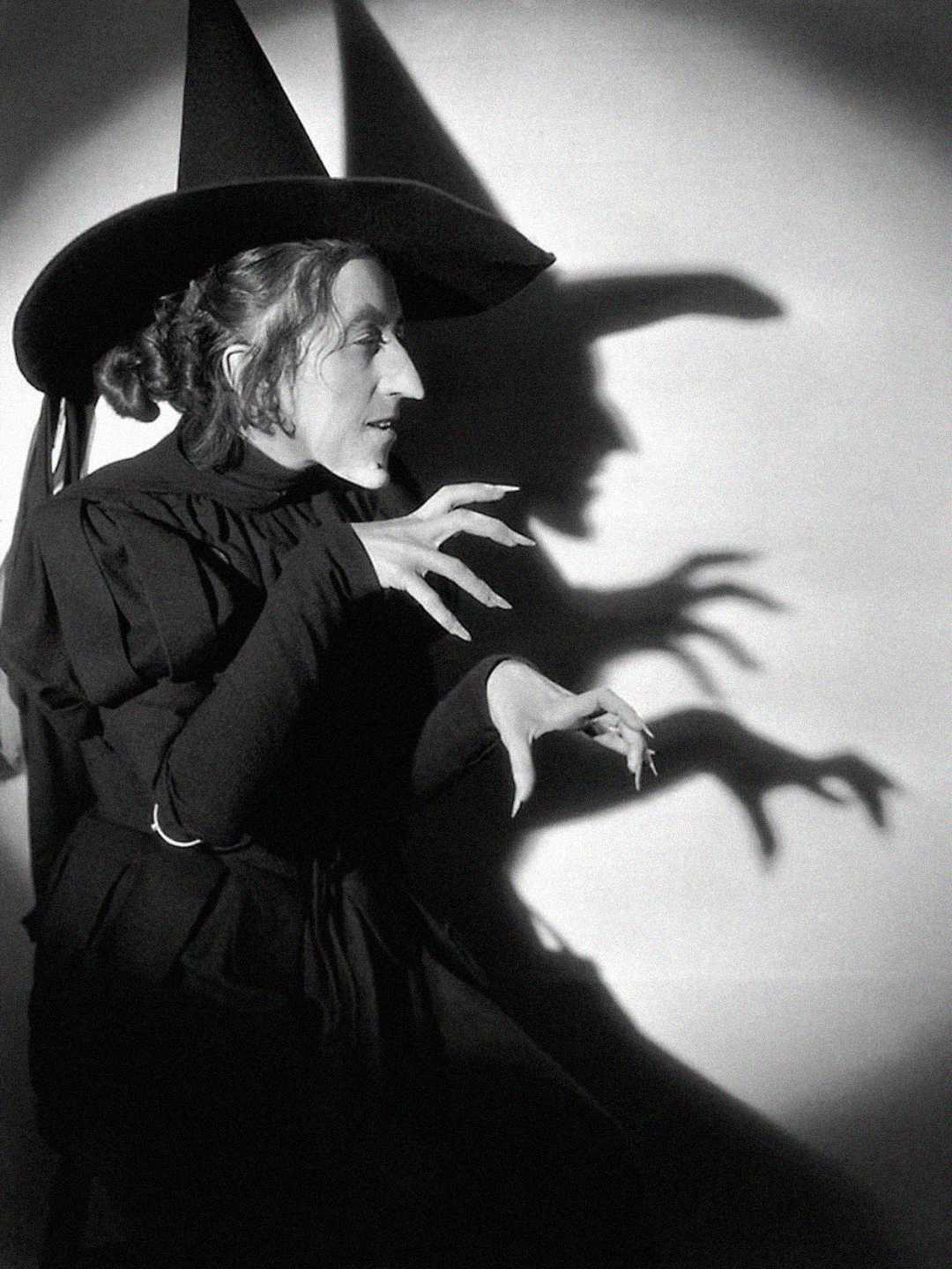 A person dressed as a witch in a pointed hat and dark clothing, with long, claw-like fingers casting dramatic shadows on the wall behind them.
