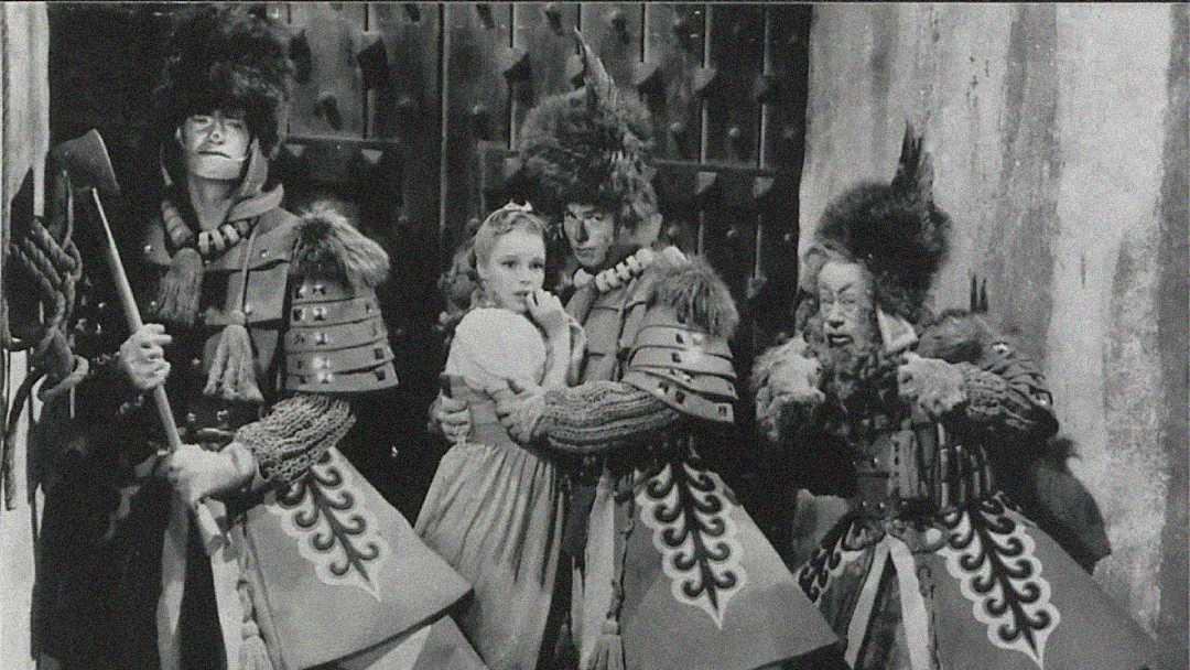 A young girl in a dress looks worried as she is surrounded by three men in elaborate, fanciful guard costumes with large feathered hats and patterned robes, standing against a stone wall.