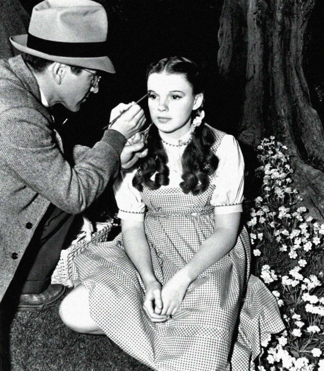 A man applies makeup to a young woman with dark curls and braided pigtails, who is seated outdoors in a gingham dress beside blooming flowers and a tree. The image is in black and white.