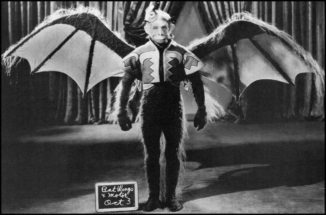 A person in a furry costume with large bat wings, a horned headpiece, and zigzag patterns on the chest stands onstage. A chalkboard at their feet reads "Bat Wings & Mask Oct 3." Curtains hang in the background.